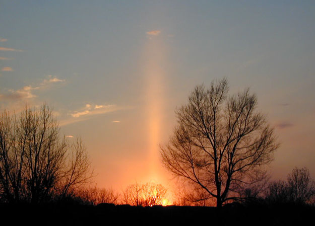 Un pilier formé par la lumière du Soleil couchant, se réflétant sur des crystaux de glace plats tombant lentement vers le sol 